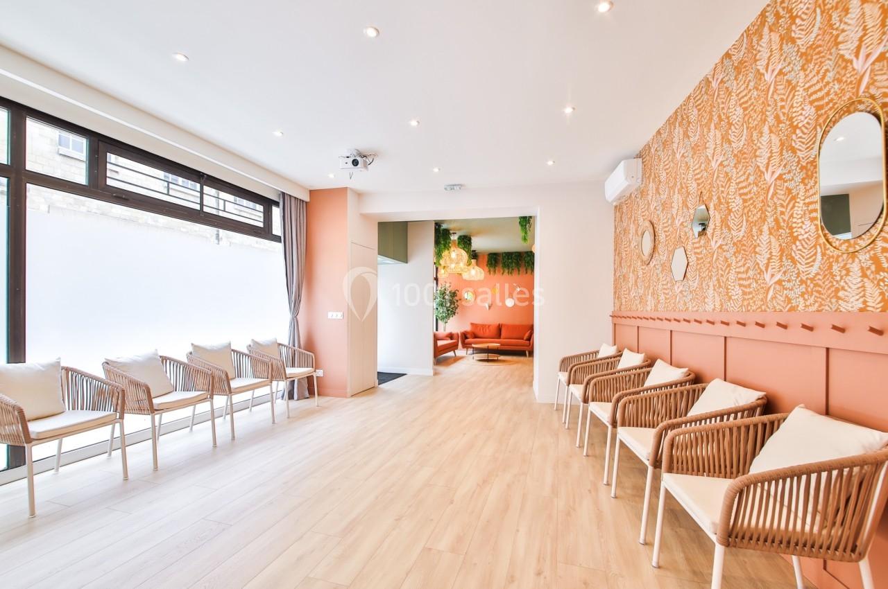 Salle d'attente lumineuse avec chaises en rotin, murs décorés de papier peint orange et grandes fenêtres.