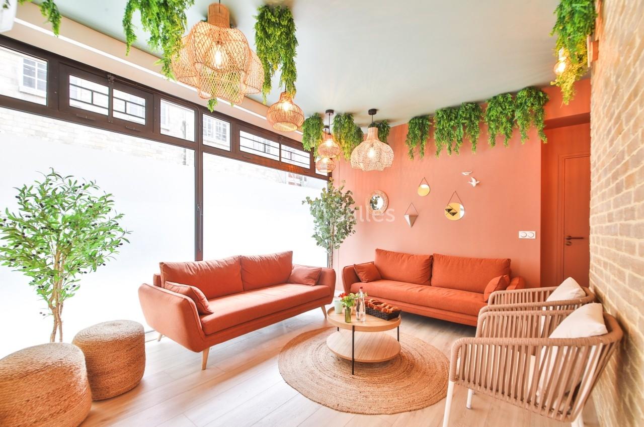 Salon lumineux avec canapés orange, plantes suspendues, murs corail et grandes fenêtres laissant entrer la lumière naturelle.