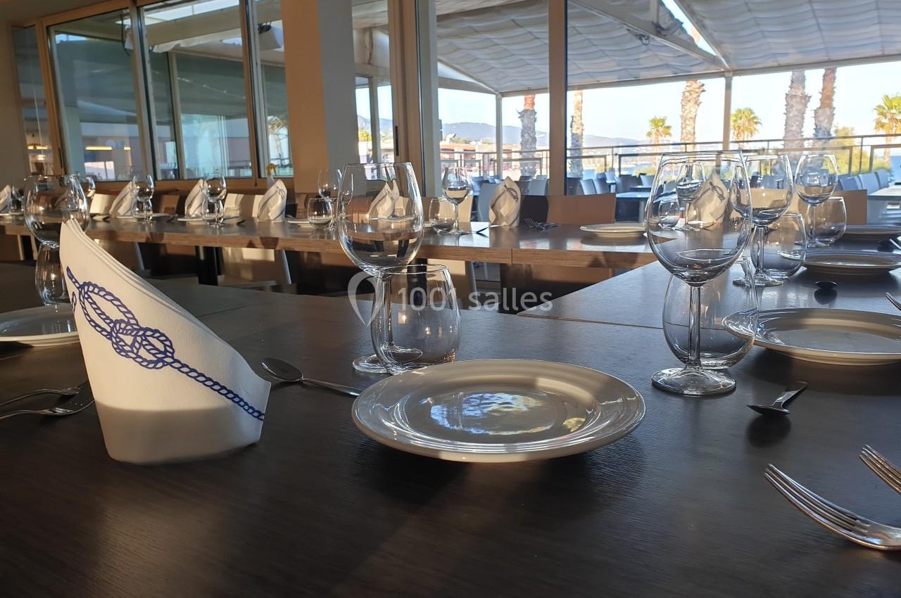 Table dressée avec assiettes, verres et serviettes pliées, dans un restaurant avec vue sur une terrasse ensoleillée.