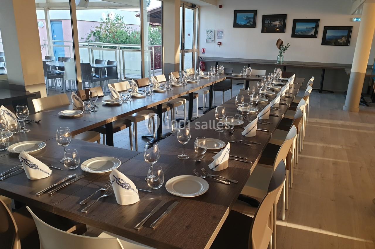 Salle lumineuse avec de grandes tables dressées, comprenant assiettes, verres et serviettes pliées, prête pour un repas.