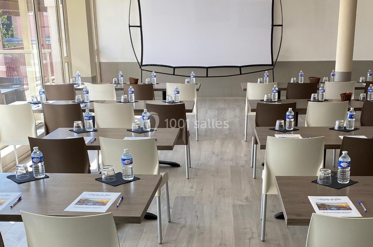 Salle de réunion lumineuse avec des tables disposées en rangées, équipées de bouteilles d'eau, stylos et documents.