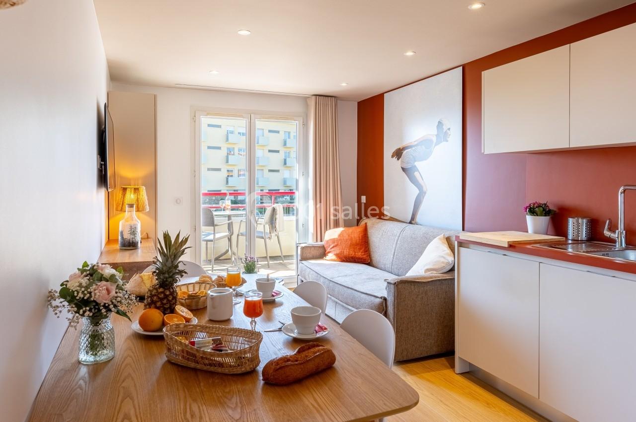 Salon avec kitchenette, table dressée pour le petit-déjeuner, canapé, et balcon donnant sur des immeubles.