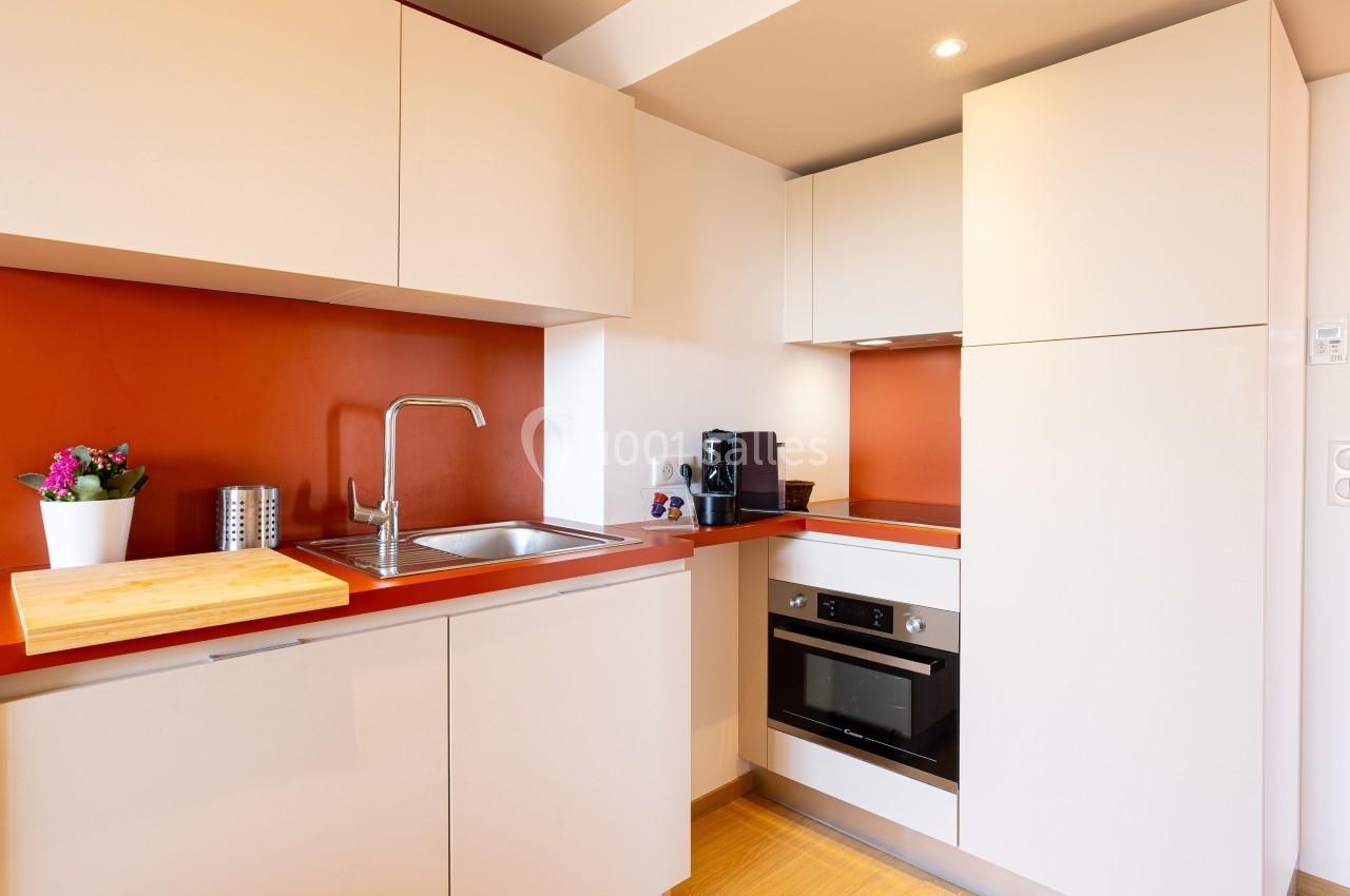 Cuisine moderne avec meubles beige, plan de travail rouge, évier inox, four encastré, et cafetière noire.