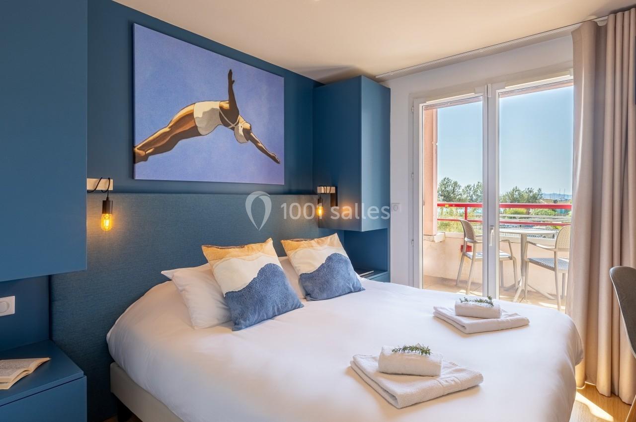 Chambre lumineuse avec lit double, décor bleu, tableau mural, et balcon donnant sur une terrasse ensoleillée.