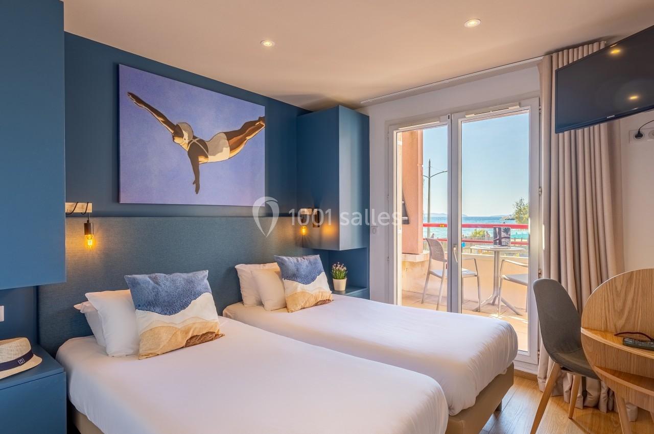 Chambre lumineuse avec deux lits simples, décoration moderne, tableau mural, balcon avec vue sur la mer et mobilier en bois.