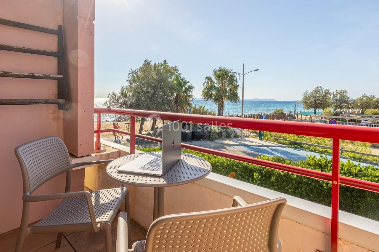 Balcon avec table, deux chaises et un ordinateur portable, vue sur la mer, des arbres et une route en arrière-plan.