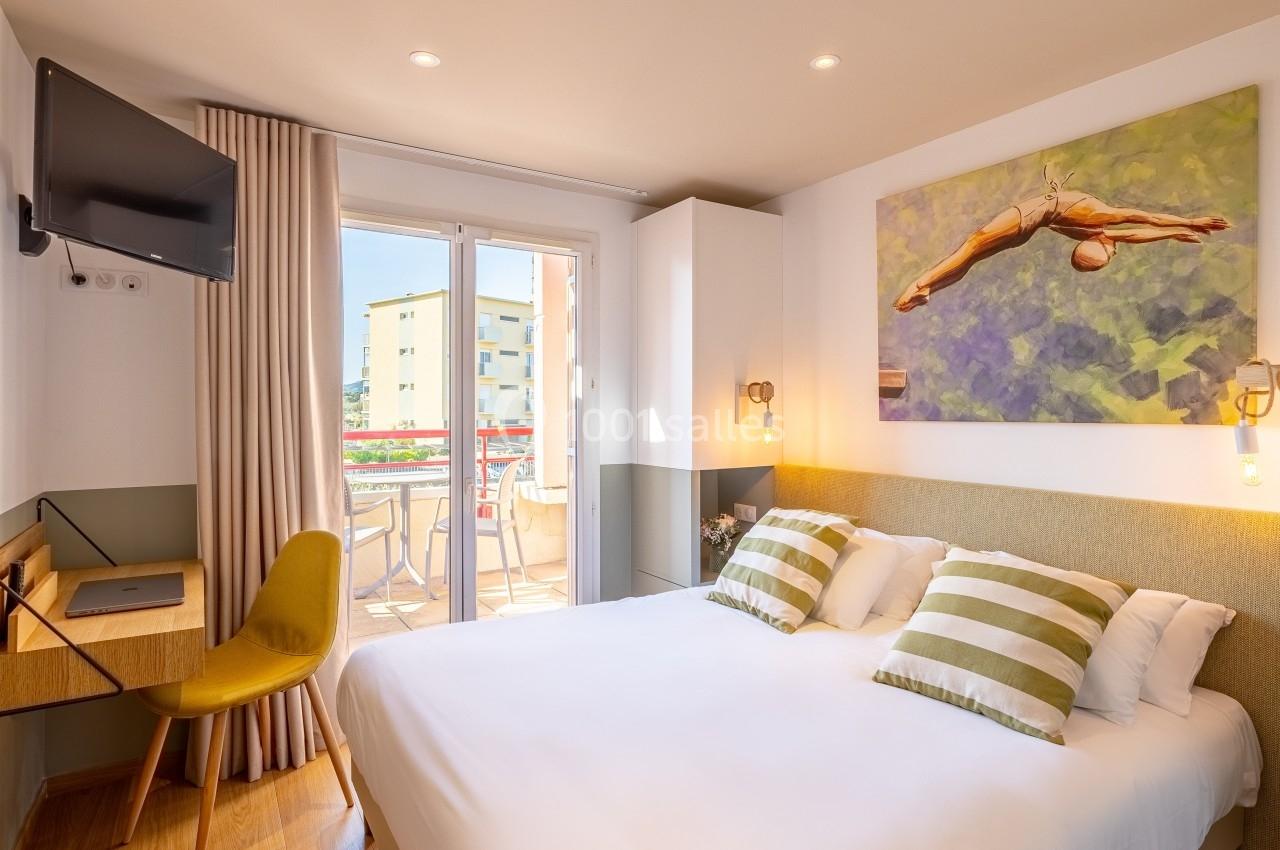 Chambre lumineuse avec lit double, bureau, télévision murale et accès à un balcon avec vue sur des bâtiments.