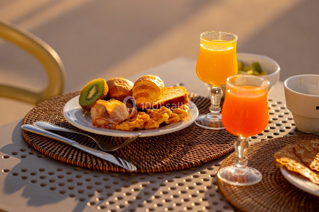 Petit-déjeuner composé de viennoiseries, fruits, œufs, jus d'orange et café, servi sur une table ensoleillée.