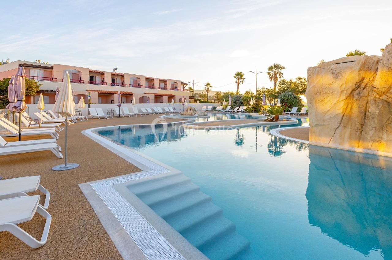 Piscine extérieure avec transats, parasols et rochers décoratifs, entourée de bâtiments et de palmiers.