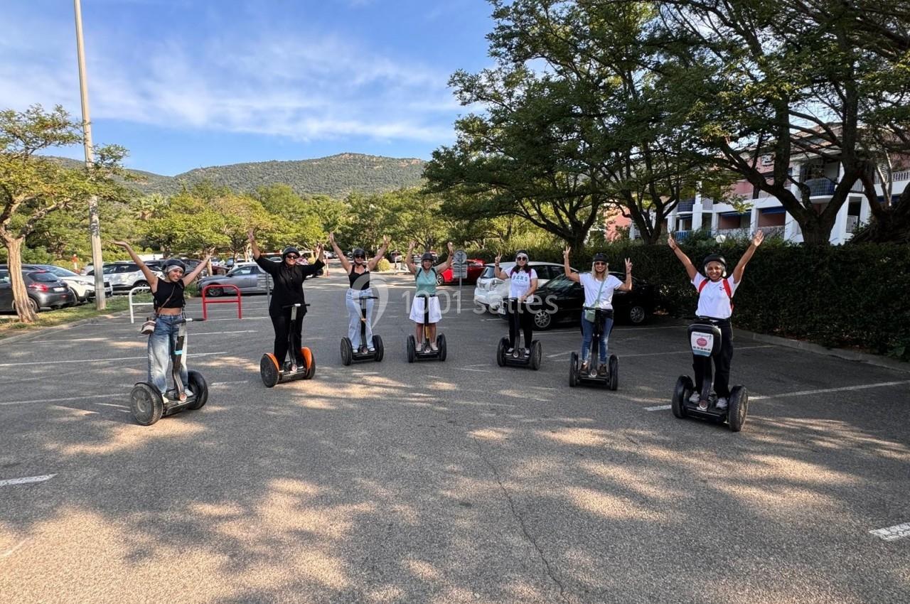 Un groupe de personnes souriantes sur des gyropodes, bras levés, dans un parking entouré d'arbres et de montagnes.