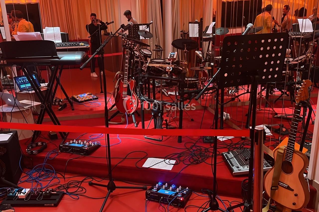 Salle de répétition avec instruments de musique, micros, câbles au sol et musiciens en arrière-plan.