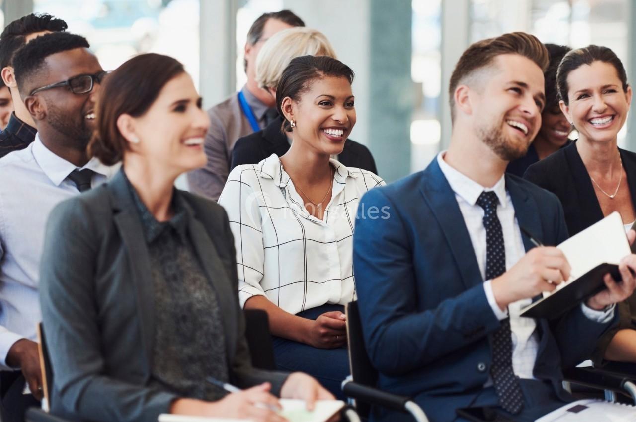 Un groupe de personnes assises dans une salle, souriant et écoutant attentivement une présentation.