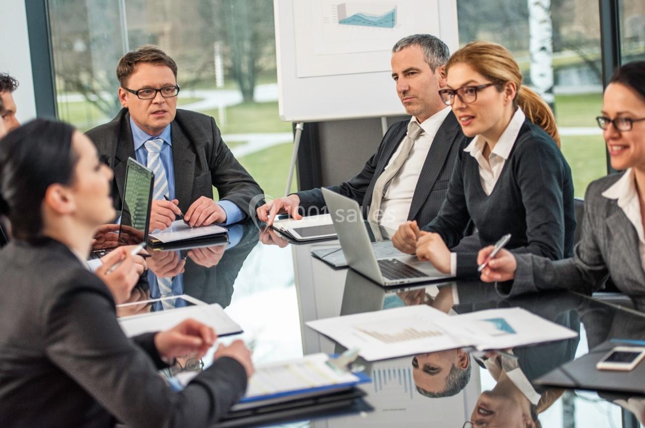 Réunion d'affaires avec plusieurs personnes en costume discutant autour d'une table dans une salle lumineuse.