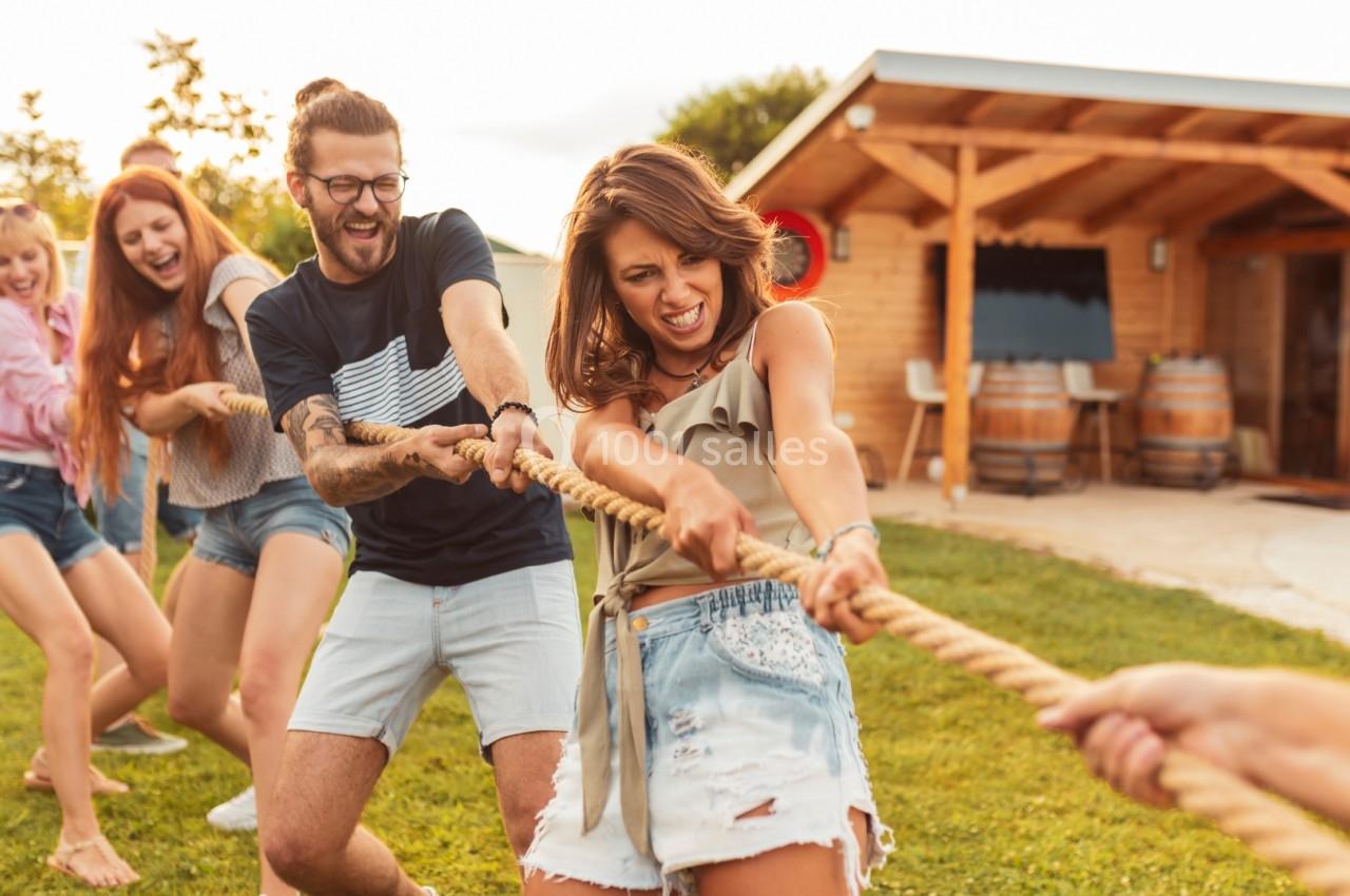 Des personnes jouent au tir à la corde dans un jardin, souriantes et concentrées, près d'une maison en bois.