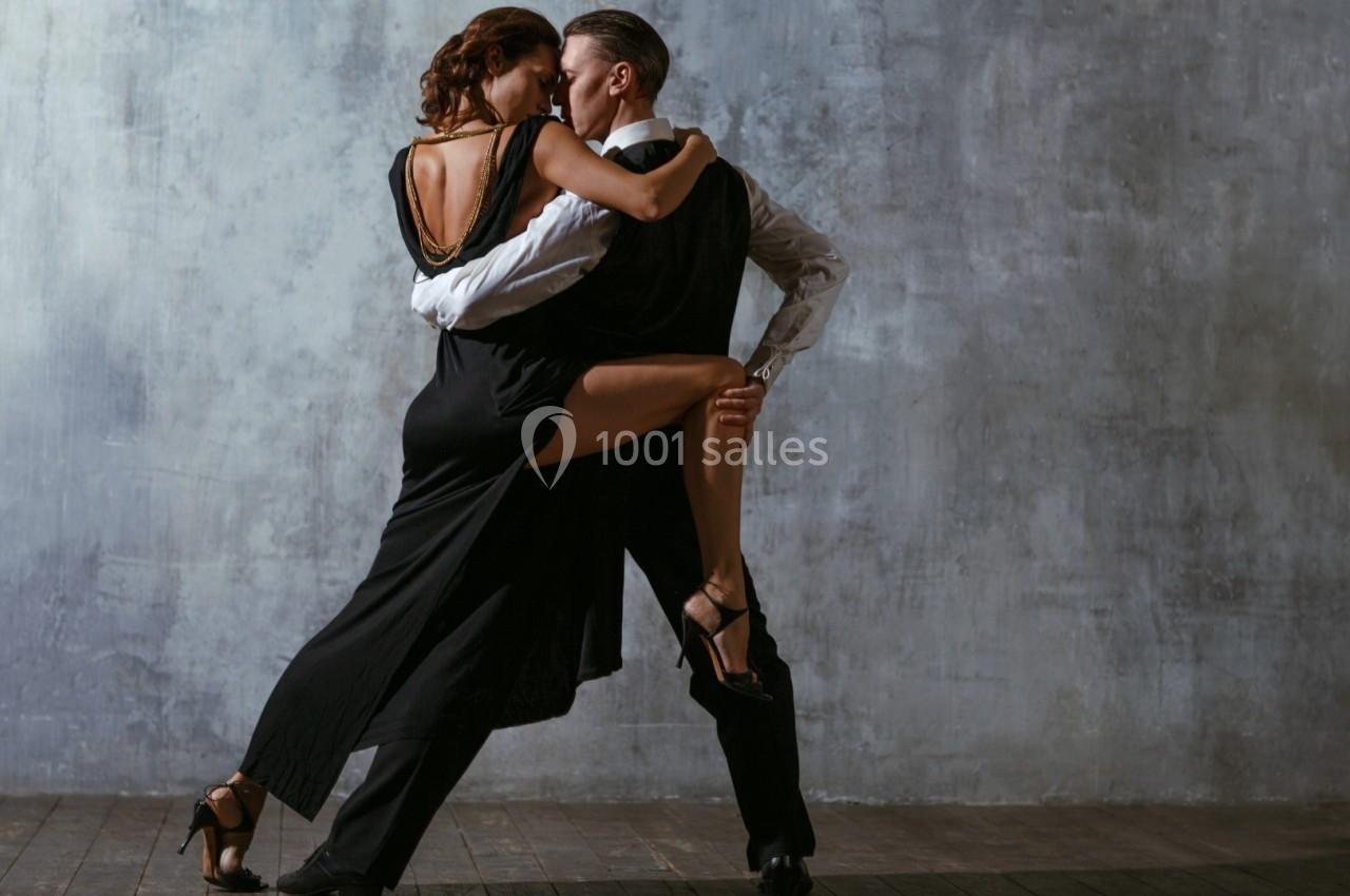 Un couple danse un tango passionné dans une salle au mur gris, éclairée par une lumière douce.