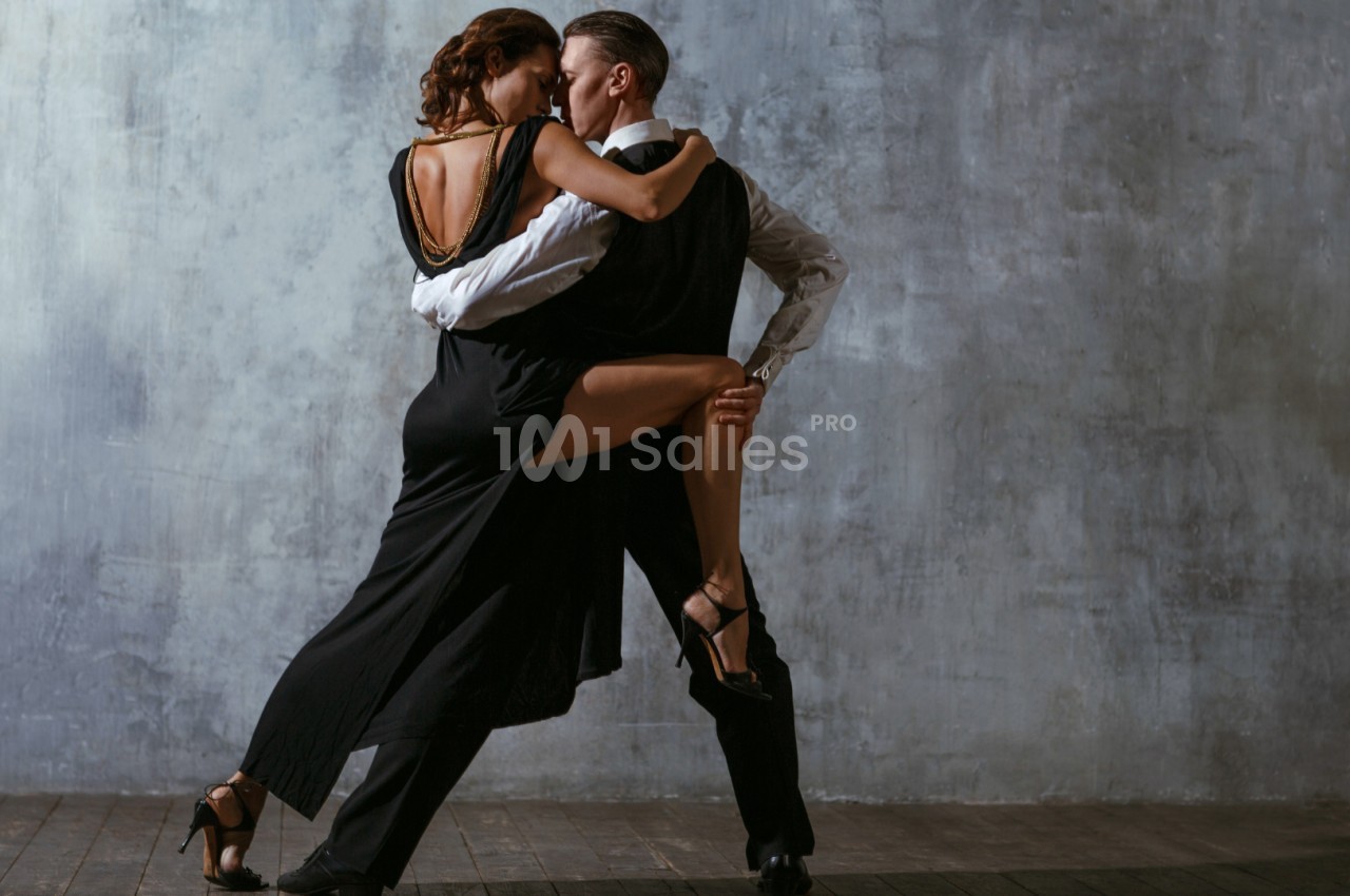 Un couple danse un tango passionné dans une salle au mur gris, éclairée par une lumière douce.