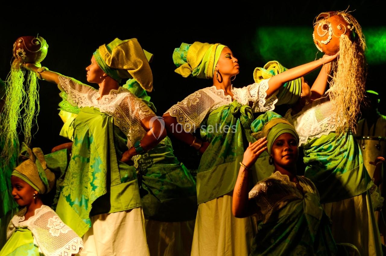 Danseuses en tenue traditionnelle verte et blanche portant des calebasses sur scène, éclairées par une lumière chaude.