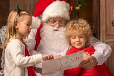 Un homme déguisé en Père Noël lit une lettre avec deux enfants souriants dans un décor chaleureux.
