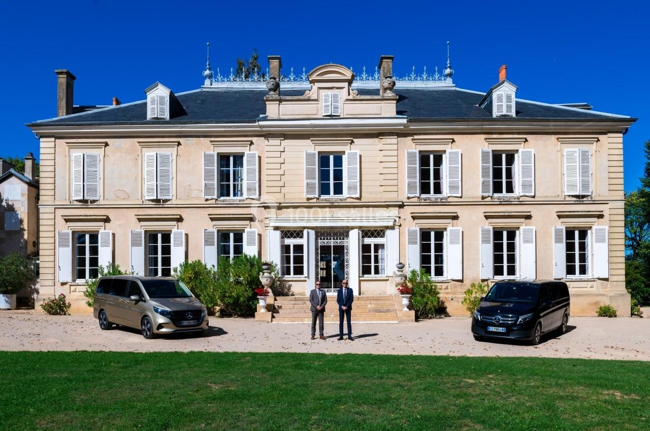 Façade d'un grand bâtiment de style classique avec des volets blancs, deux véhicules noirs et deux personnes devant.