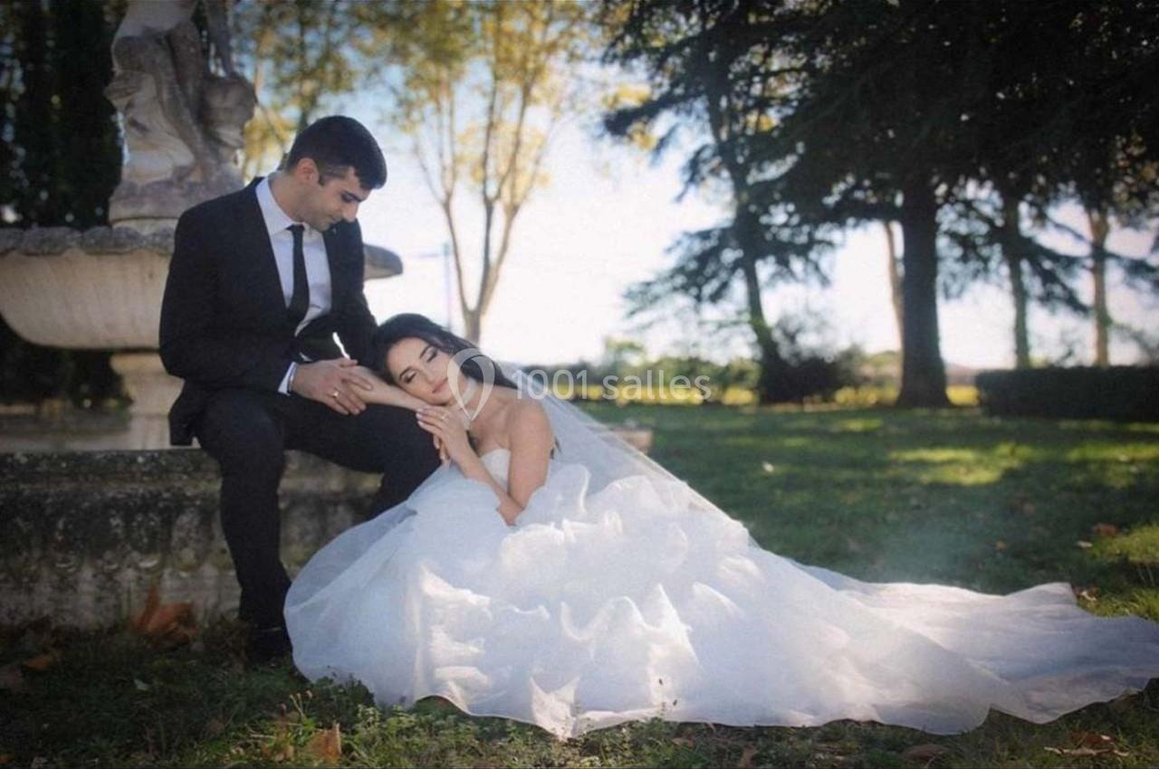 Un couple en tenue de mariage, assis près d'une fontaine dans un parc verdoyant sous un ciel ensoleillé.