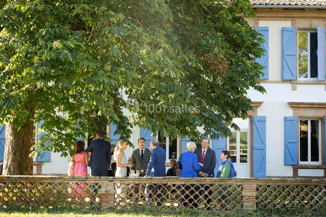 Groupe de personnes discutant sous un grand arbre devant une maison blanche aux volets bleus.