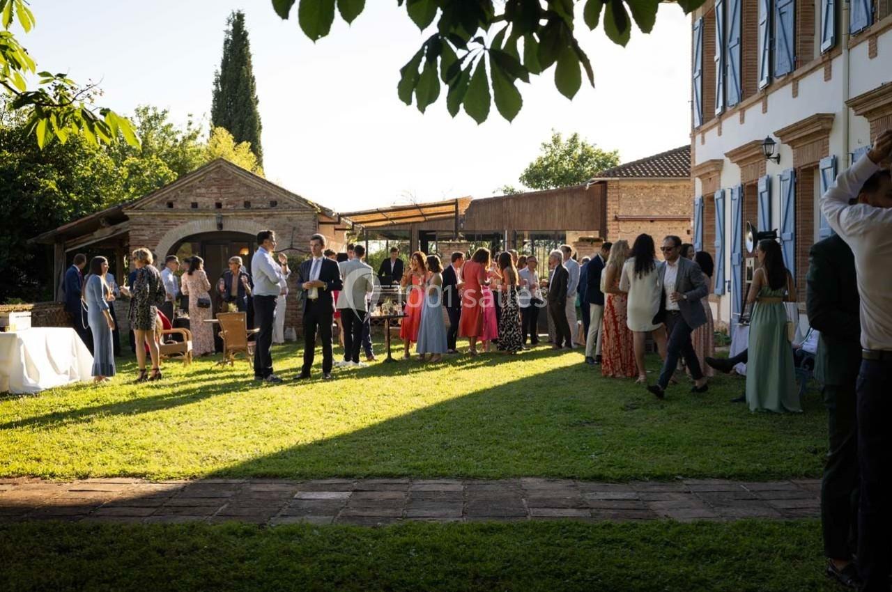 Groupe de personnes discutant dans un jardin ensoleillé, près d'une maison et d'une dépendance en pierre.
