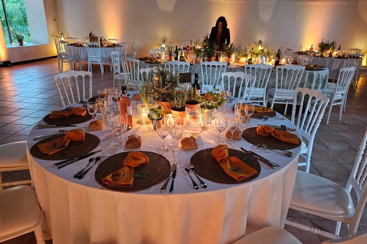 Salle de réception décorée avec des tables rondes dressées, nappes blanches, serviettes orange et lumière tamisée.