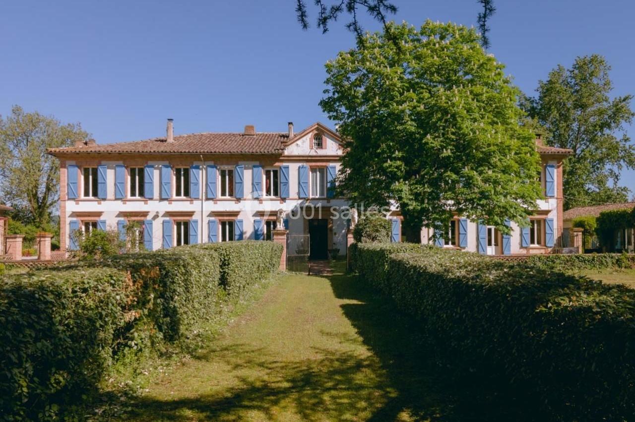 Grande maison en briques avec volets bleus, entourée de haies et d'arbres, sous un ciel dégagé.