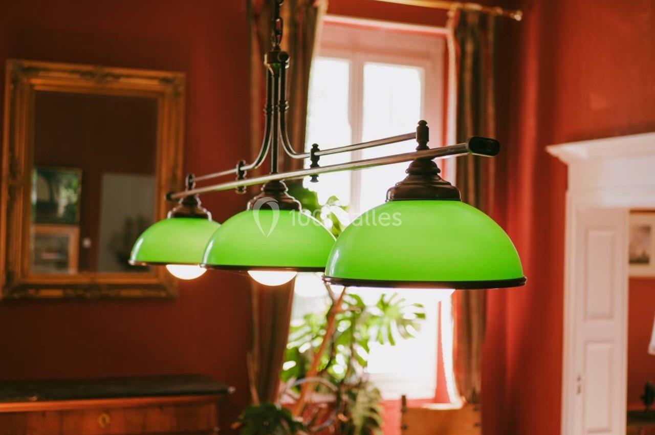 Suspension avec abat-jours verts dans une pièce aux murs rouges, éclairée par une fenêtre en arrière-plan.