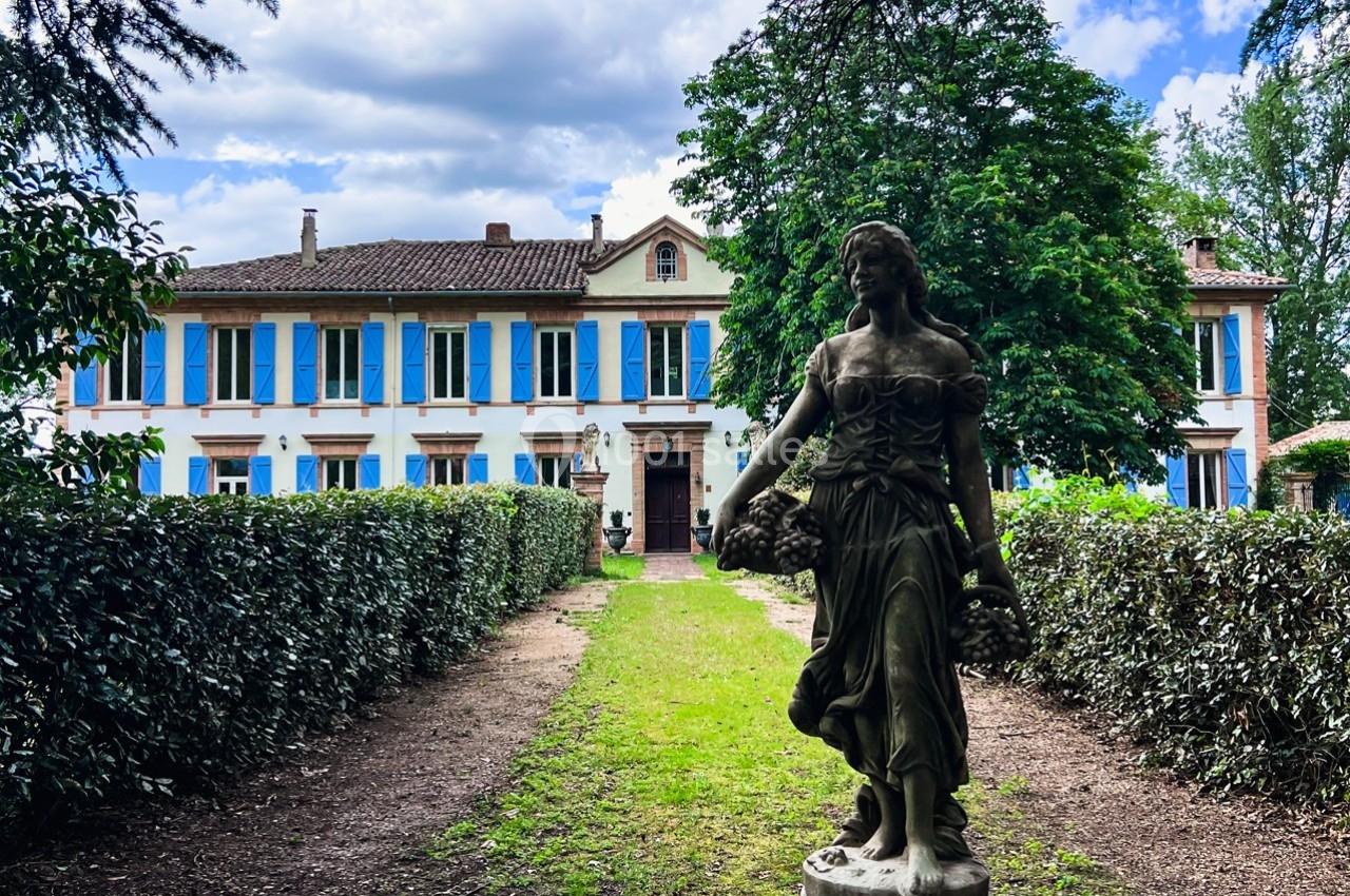 Statue en pierre d'une femme tenant des paniers de fleurs, devant une grande maison aux volets bleus entourée de verdure.