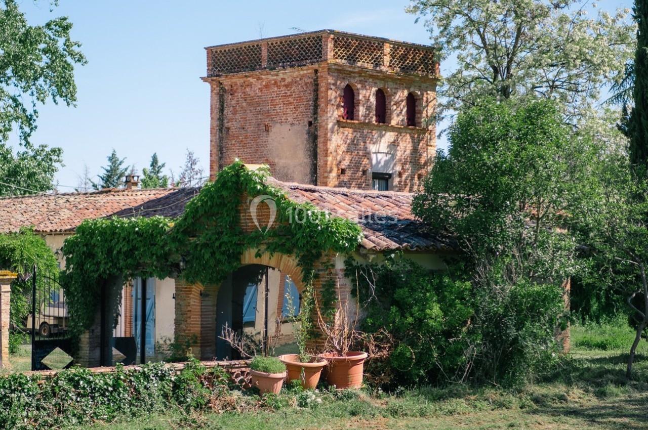 Bâtiment en briques avec une tour, entouré de végétation et de pots de fleurs sur une pelouse.