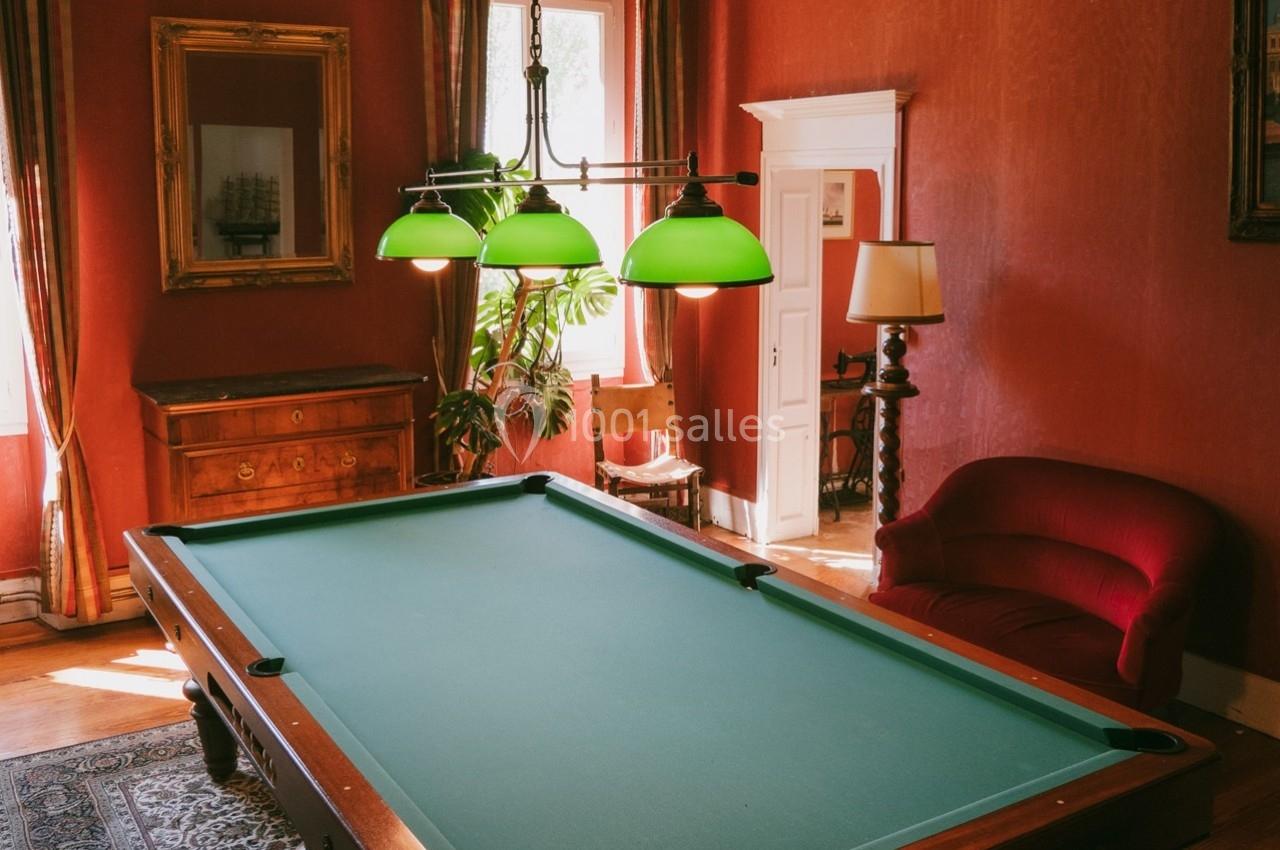 Table de billard dans une pièce aux murs rouges, éclairée par une suspension à trois lampes vertes.