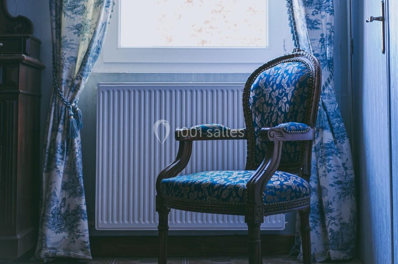 Fauteuil en bois sculpté avec revêtement bleu à motifs, placé près d'une fenêtre avec rideaux assortis.