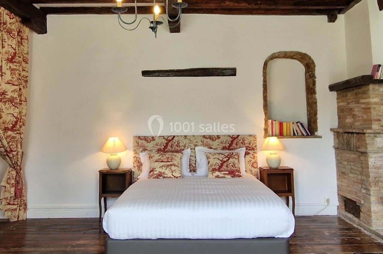 Chambre avec lit double, poutres apparentes, cheminée en briques, lampes de chevet et rideaux à motifs rouges.