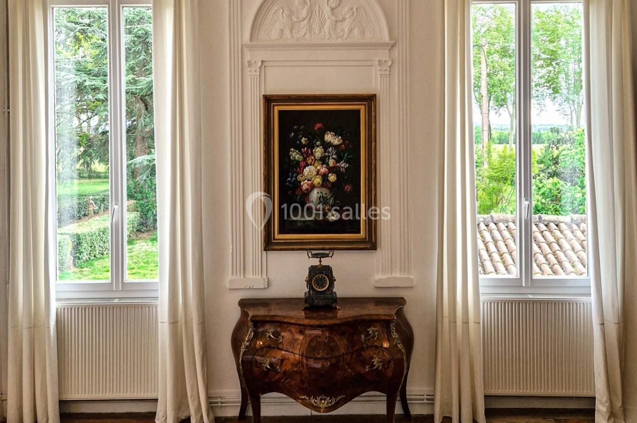 Commode en bois sculpté avec un téléphone ancien, encadrée par un tableau floral et deux fenêtres donnant sur un jardin.