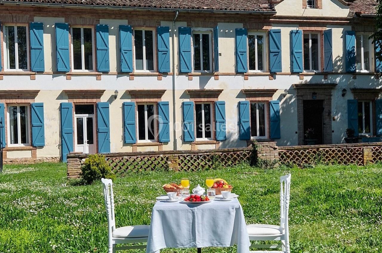 Table dressée pour un petit-déjeuner en plein air devant une grande maison aux volets bleus, entourée de verdure.