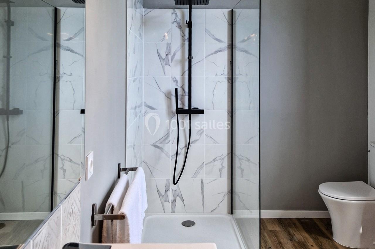 Douche moderne avec paroi vitrée, carrelage effet marbre, robinetterie noire et toilettes en arrière-plan.