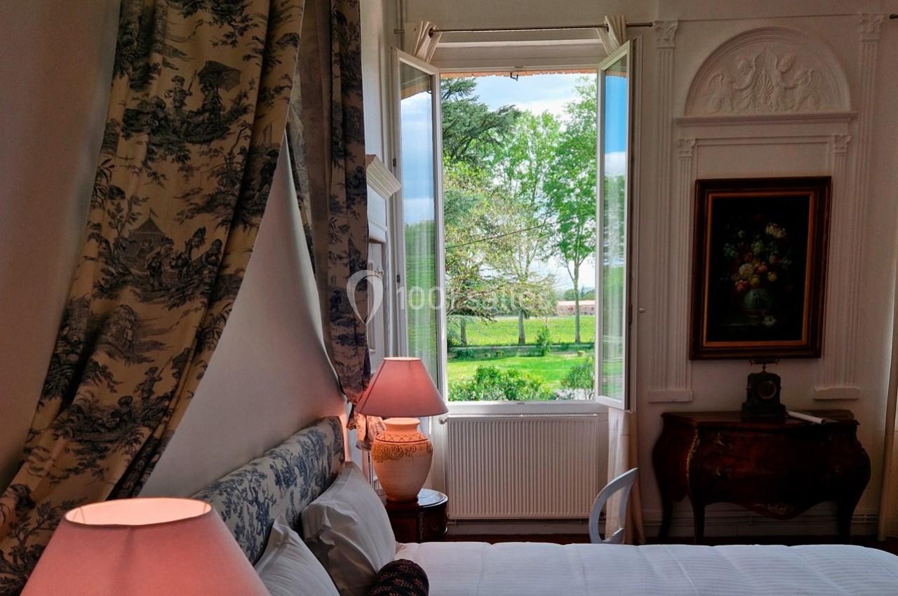 Chambre lumineuse avec lit, lampes de chevet et fenêtre ouverte offrant une vue sur un jardin verdoyant.