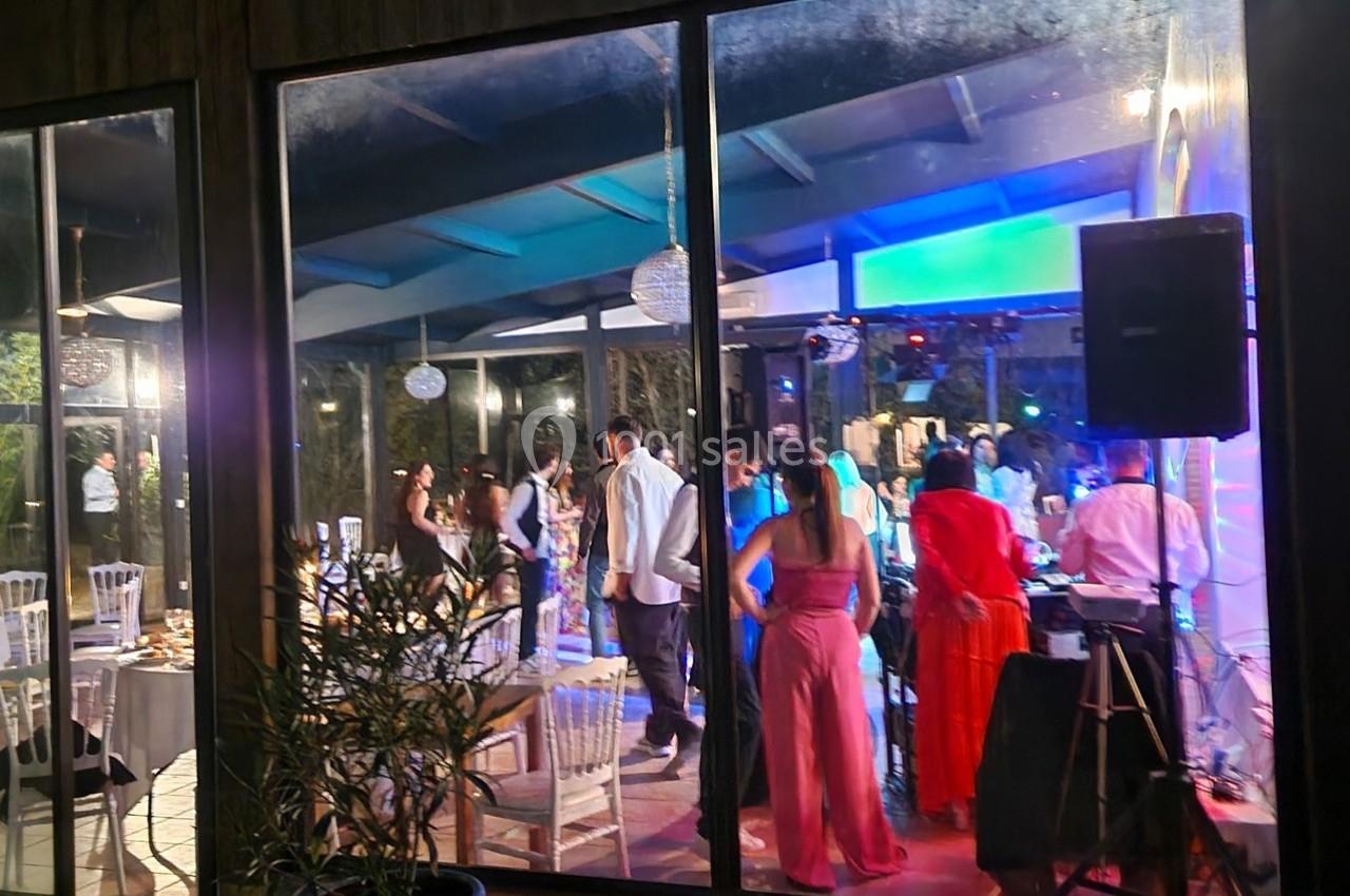 Des personnes dansent et discutent dans une salle éclairée, vue depuis l'extérieur à travers des baies vitrées.