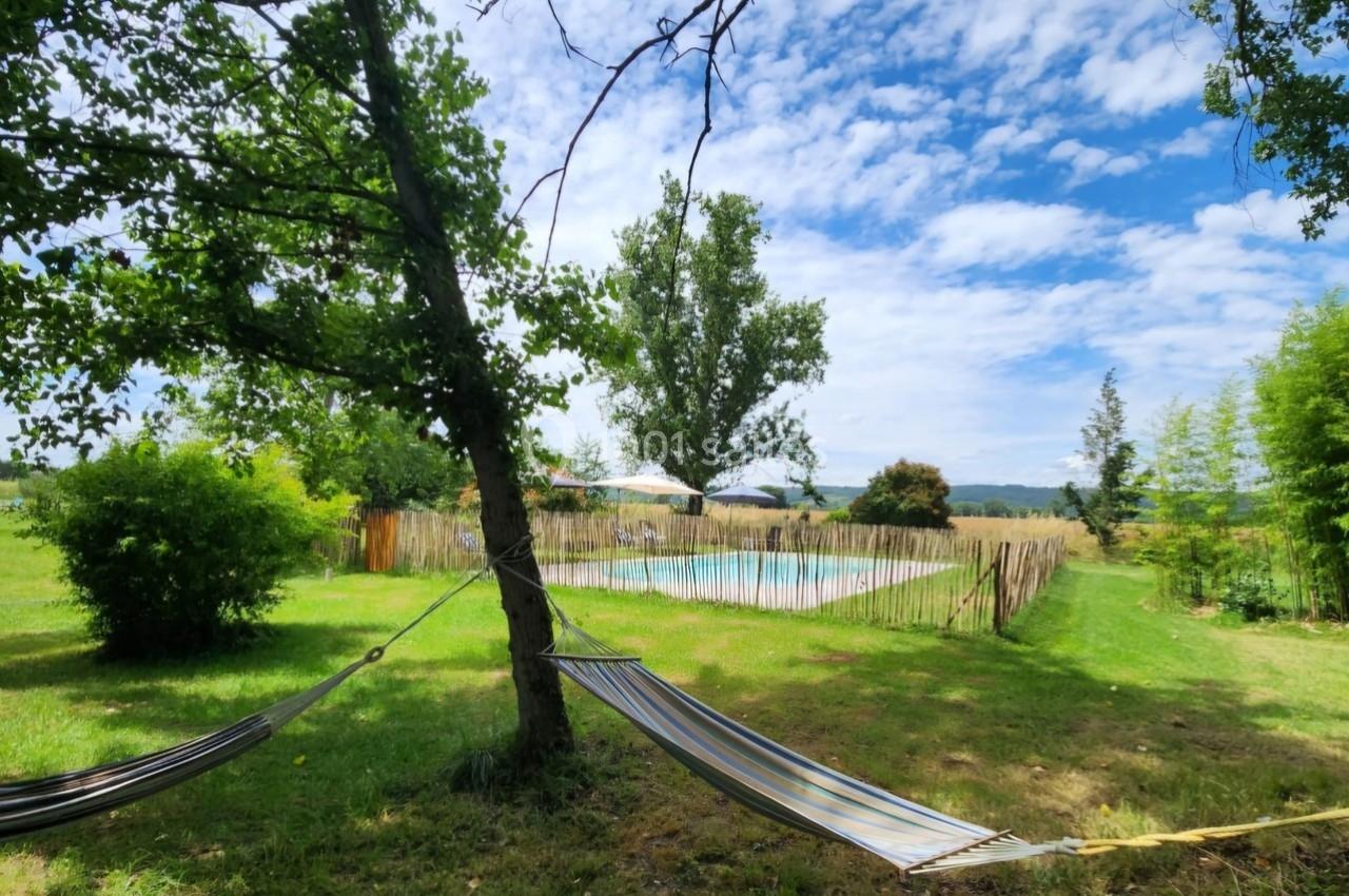 Piscine extérieure entourée de verdure et clôturée, avec des hamacs suspendus à l'ombre d'arbres.