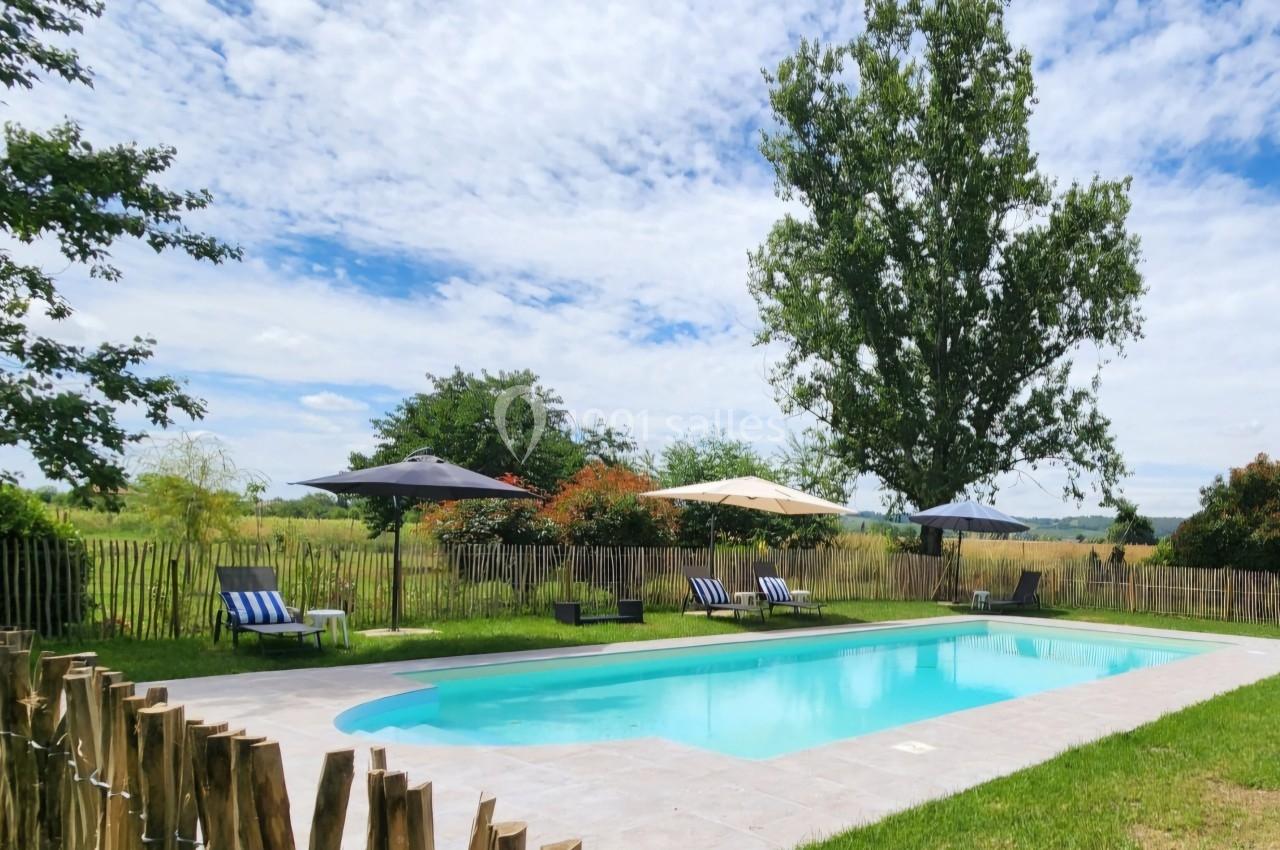 Piscine extérieure entourée de chaises longues, parasols et végétation, avec un grand arbre en arrière-plan.