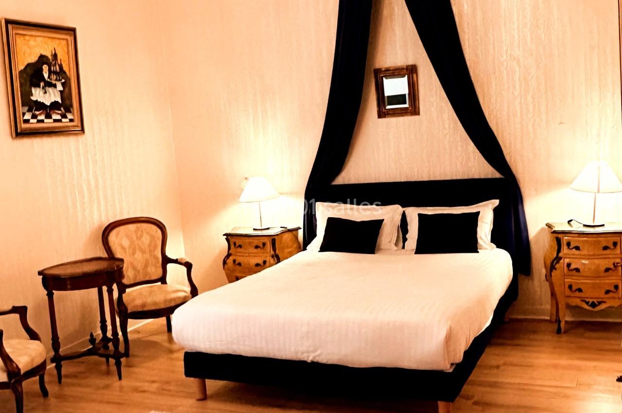 Chambre lumineuse avec lit double, baldaquin noir, meubles anciens, parquet clair et lustre décoratif.