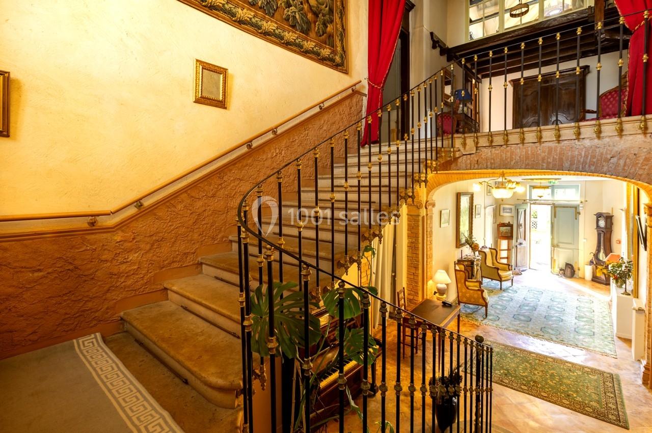 Escalier en colimaçon avec rampe en fer forgé dans un intérieur lumineux, décoré de tapis et de tableaux.