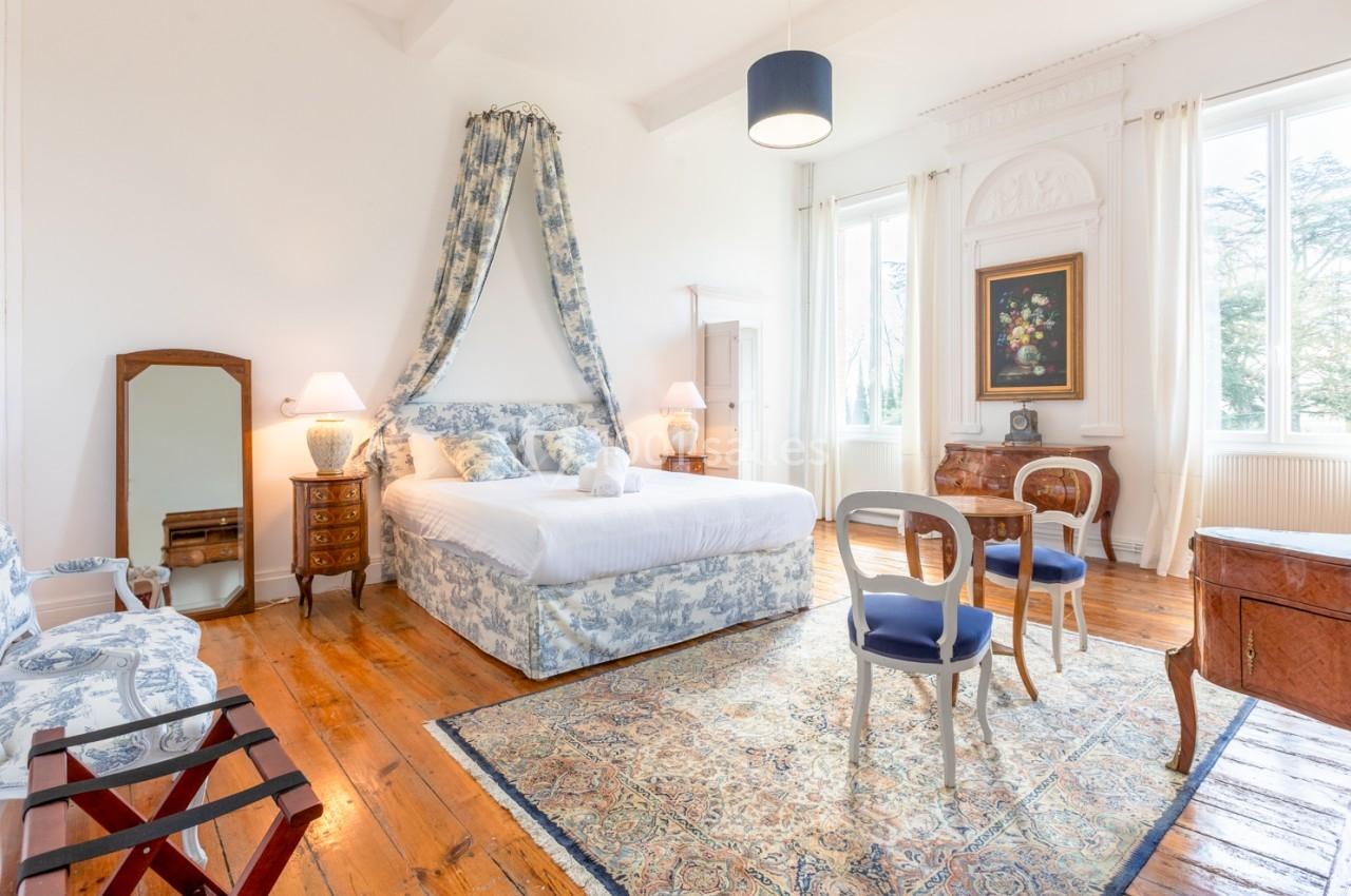 Chambre lumineuse avec lit à baldaquin, meubles anciens, tapis décoratif et grandes fenêtres donnant sur l'extérieur.