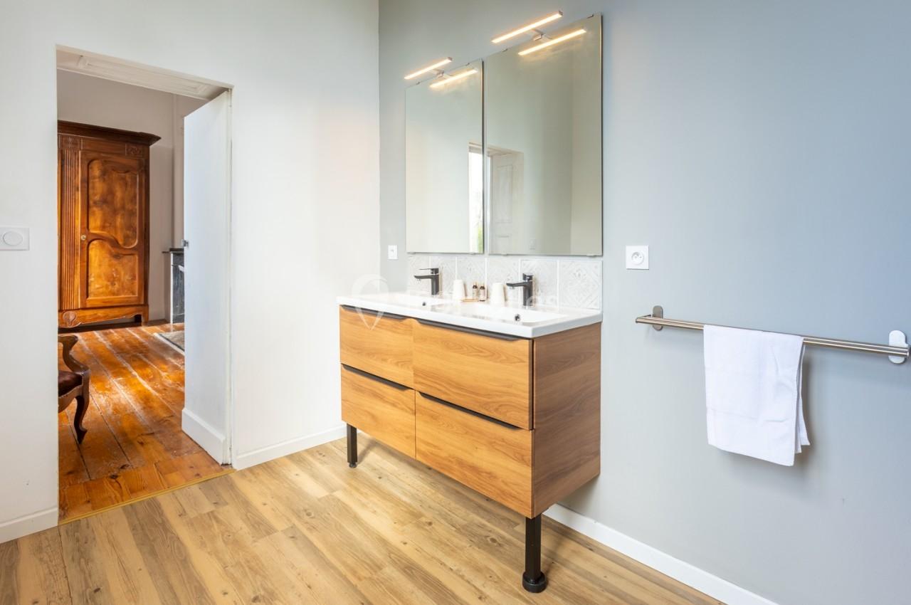 Salle de bain moderne avec meuble en bois, double vasque, miroir éclairé et porte ouverte sur une pièce avec parquet.