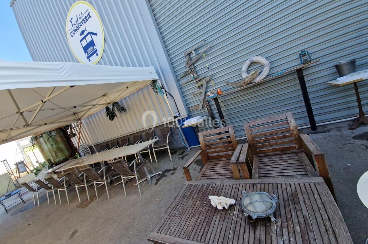 Mobilier en bois et tables sous un auvent près d'un bâtiment métallique avec un logo circulaire visible.