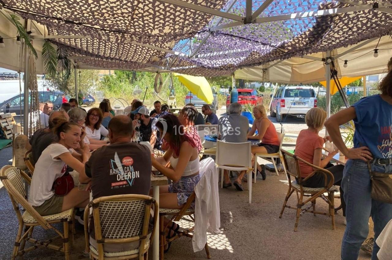 Des personnes assises à des tables sous une pergola ombragée, discutant et partageant un moment convivial.