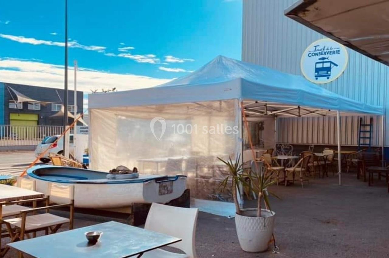 Terrasse extérieure avec tables, chaises, une barque décorative et un auvent blanc près d'un bâtiment industriel.