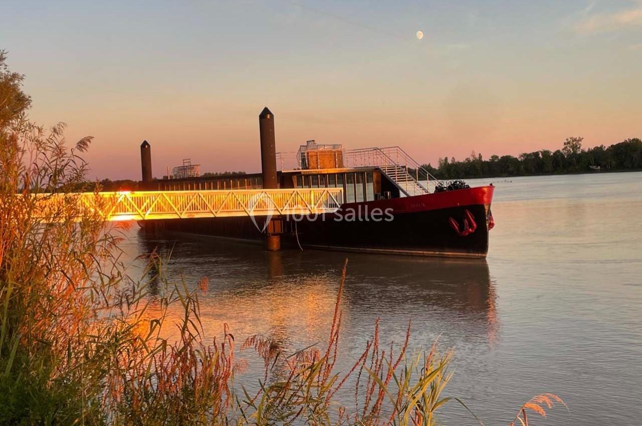Bateau amarré sur une rivière au coucher du soleil, avec une passerelle illuminée et des roseaux au premier plan.