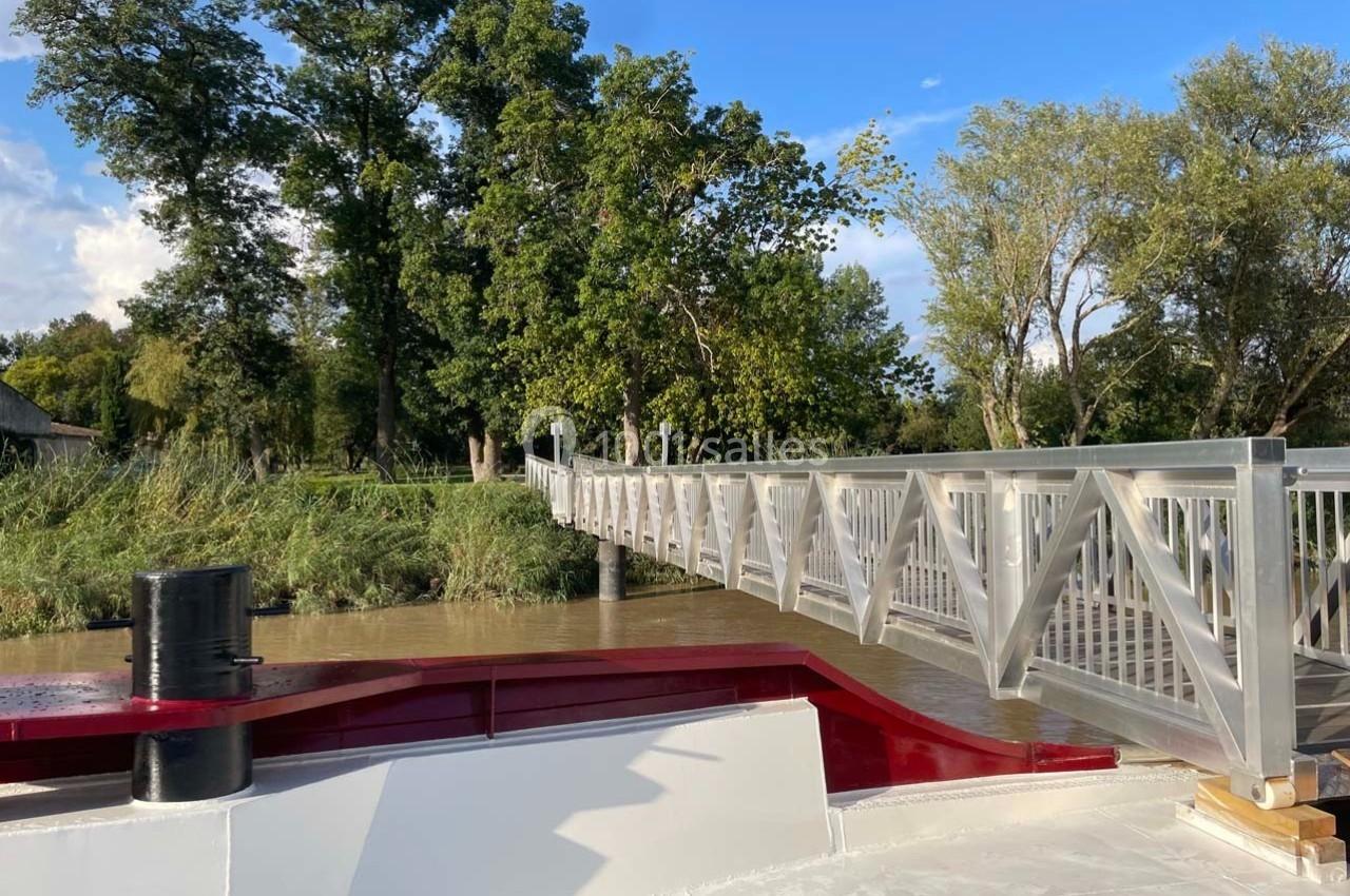Vue d'une passerelle en bois clair traversant une rivière, entourée d'arbres et d'un ciel partiellement dégagé.